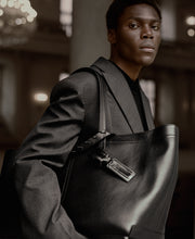Load image into Gallery viewer, RT_XL Reinstote XL in black color, showing mood picture of model wearing the bag from aside. It also shows the Luggage Tag Eel at the handle of the bag.